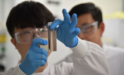 A close up of gloved hands holding a magnet against a small vial of brown liquid with two faces in the background